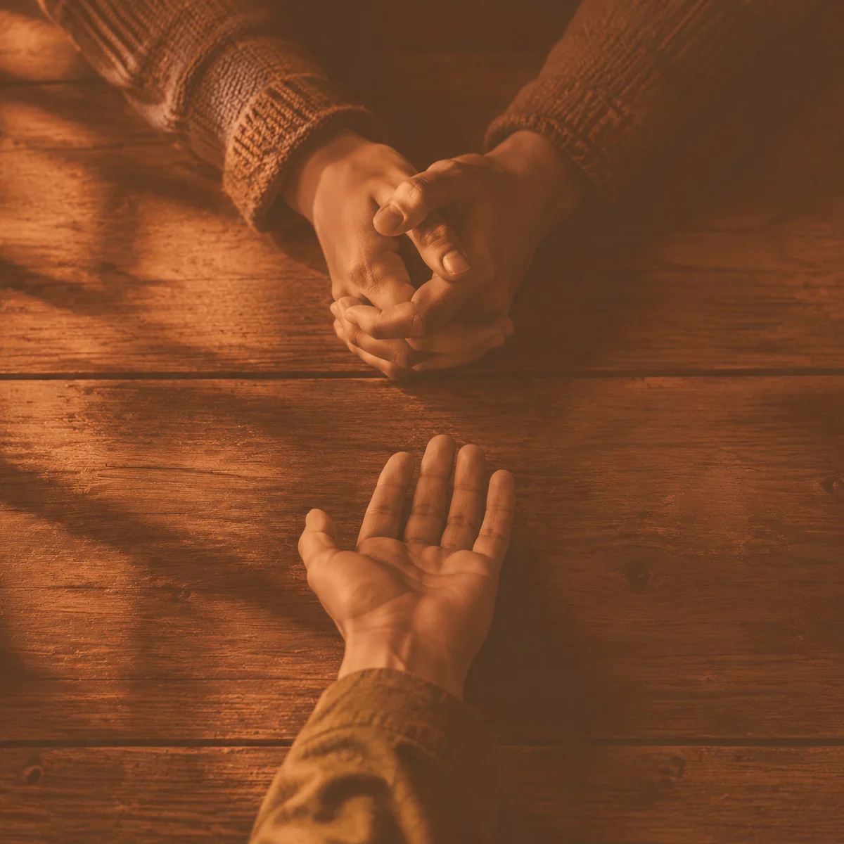 Warm-toned duotone photograph of two hands close together on a table with a third hand reaching from a distance