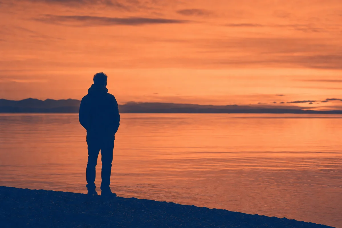 Single figure looking out across calm water in warm peach and midnight blue duotone