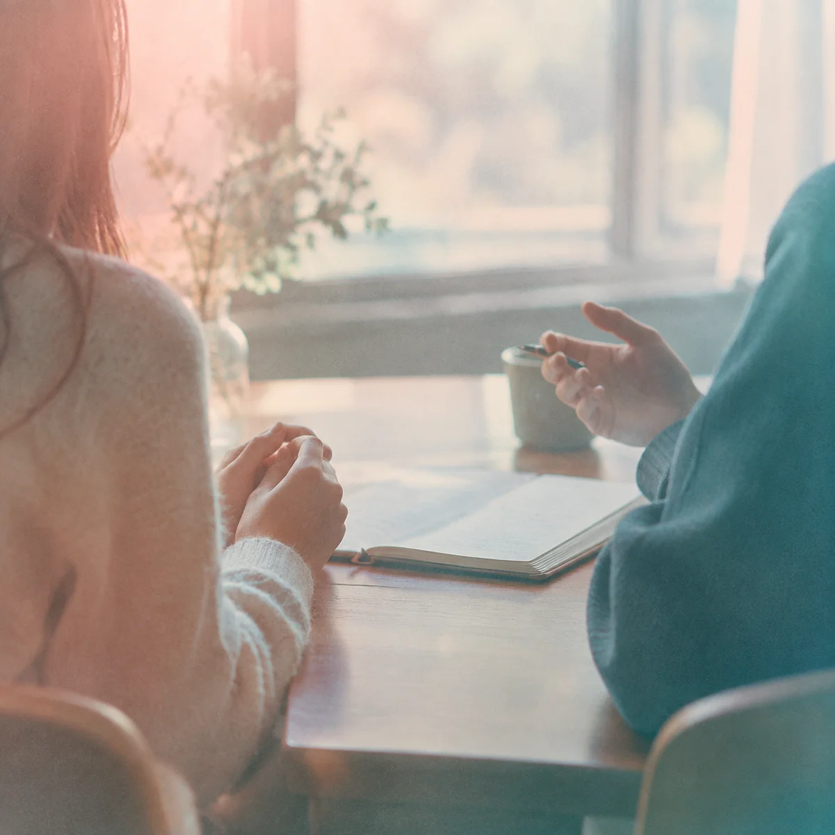 Two people seen from behind across a table, hands visible and a notebook open between them, pastel editorial photography